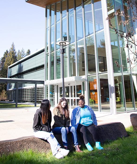 Three students sit on large rock in the sun neaby a modern glass building