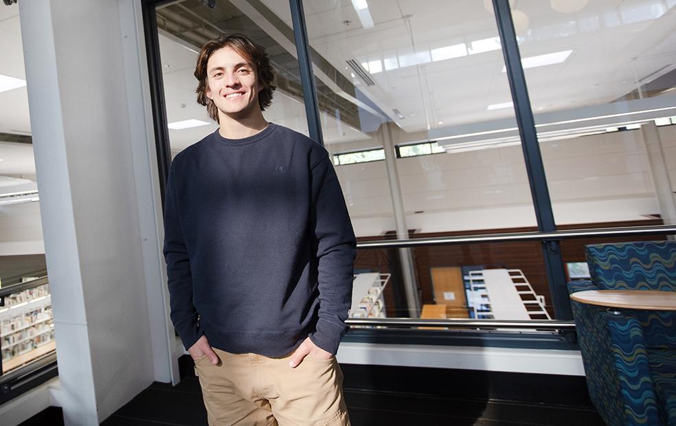 Smiling student in sweatshit stands in front of windows with hands in pants pockets