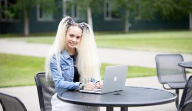 Student facing the camera smiling with two long blonde pigtails, wearing a jean jacket, typing on a computer
