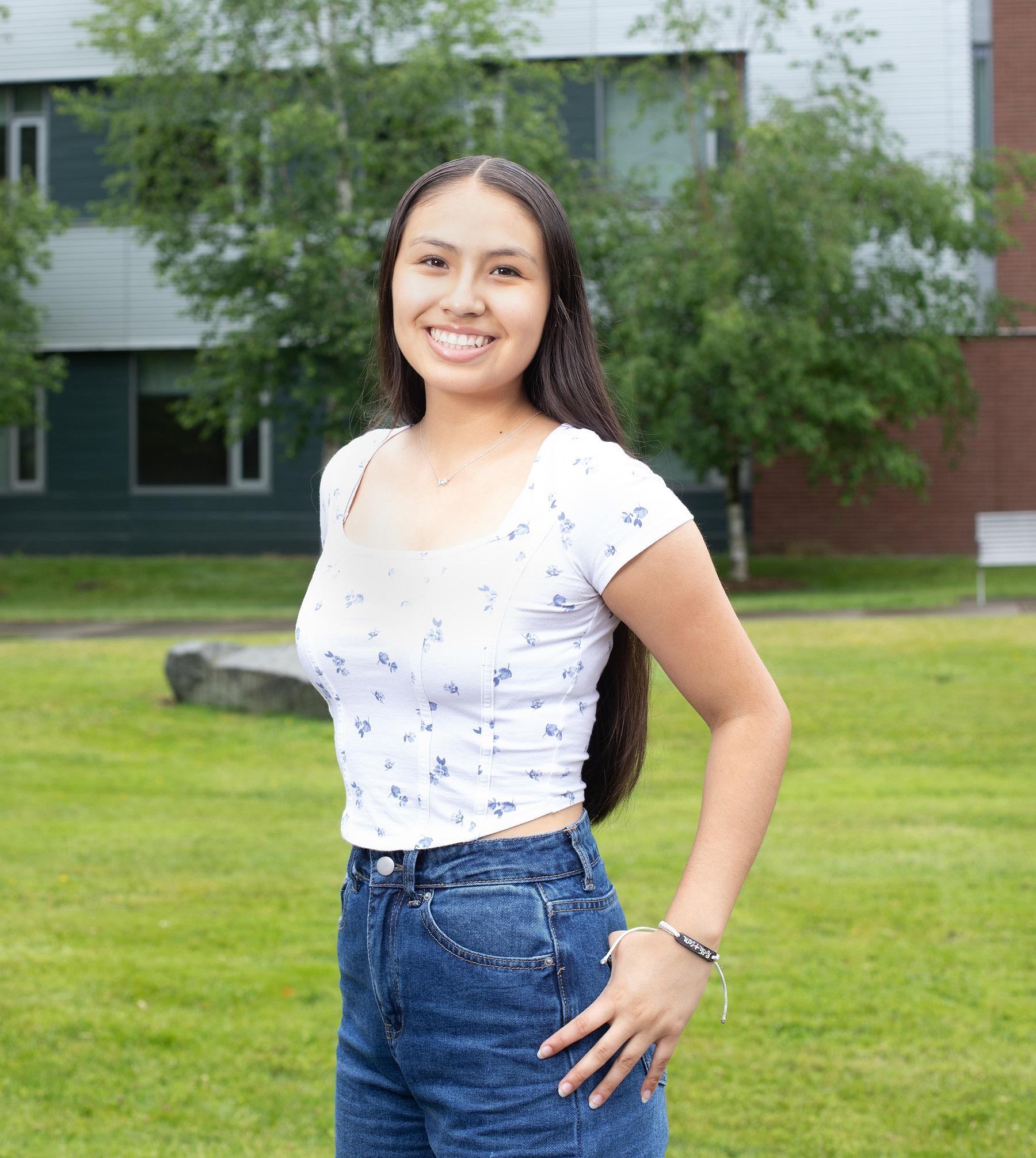 Student smiles in front of grass and building with trees in front of it