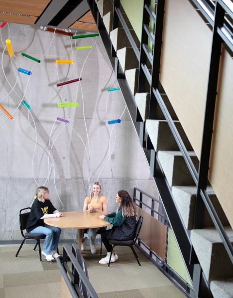 Three students sit at a round table in modern building with colorful art hanging above them