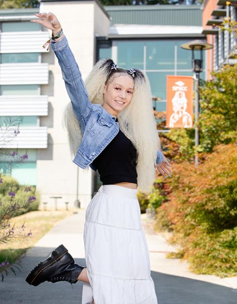 Student stands on one foot with arms in the air, smiling, outside of modern building