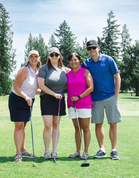 Three women and one man on a green grass holding golf clubs smiling.