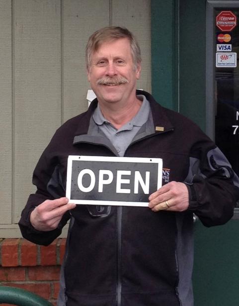 A man wearing a black jacket smiling at the camera holding a sign that reads "Open" standing in front of a building.
