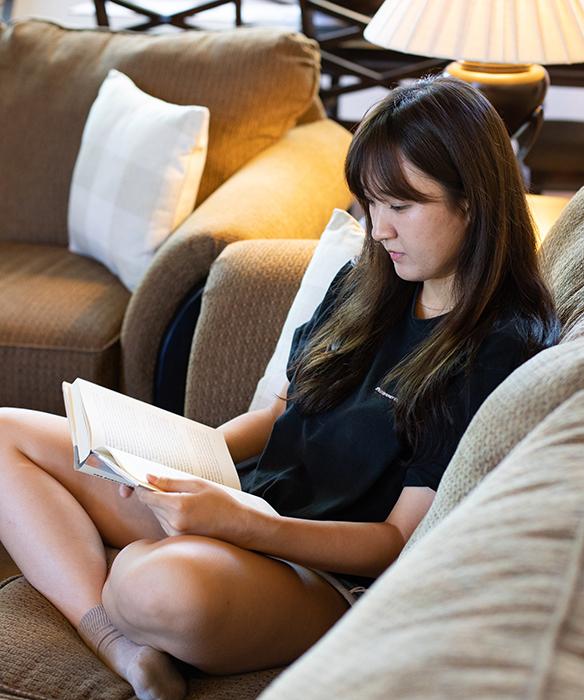 Student sitting cross-legged on a brown sofa reading a book