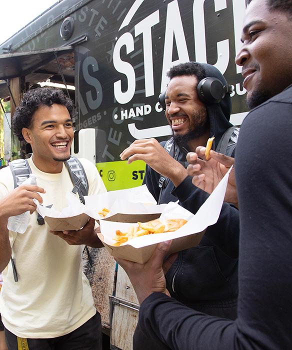 Three students laughing in conversation in front a food truck, holding to-go plates of food
