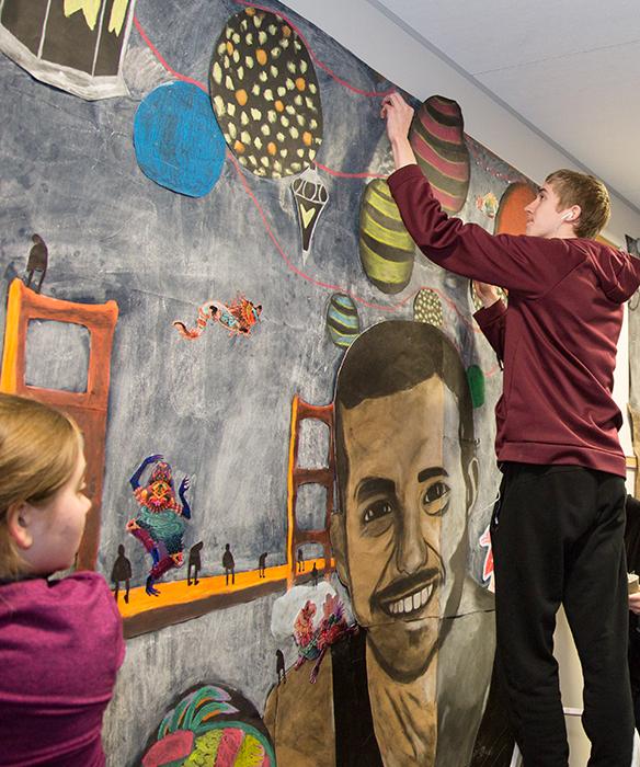 Student installs large mixed media piece on wall, standing on ladder