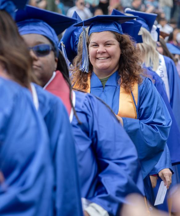 A person wearing blue graduation cap and gown smiling at the camera amid a crowd of other students.