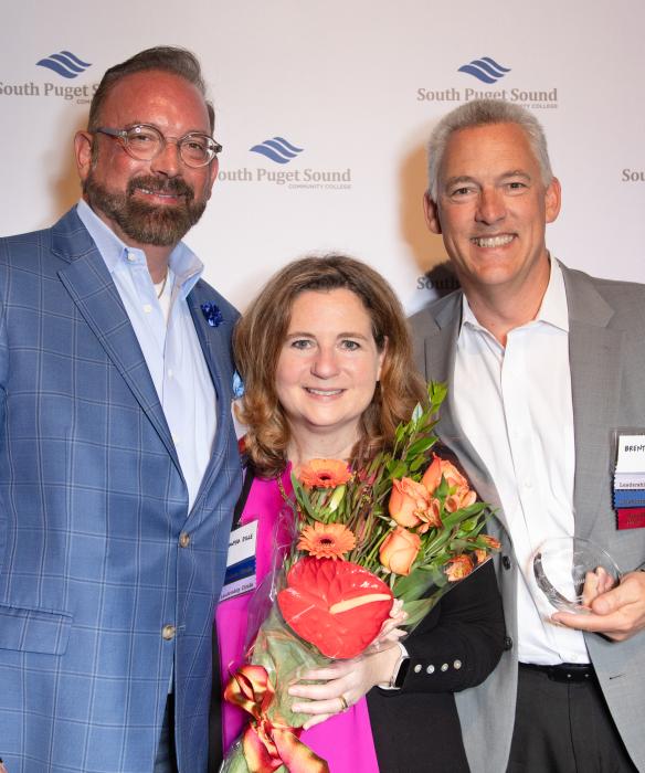 Three people looking at the camera smiling. One is holding flowers the other a glass trophy.