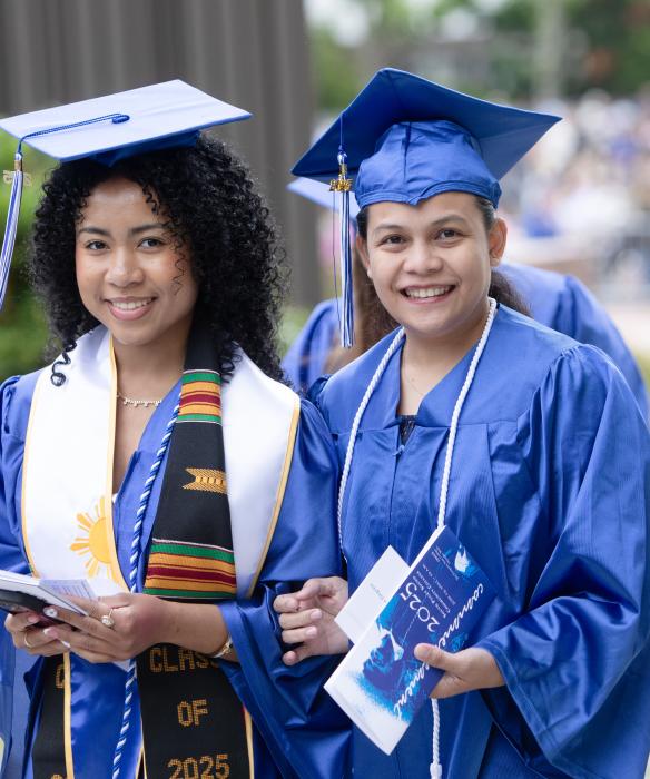 Two students in blue graduation cap and gowns smile at the camera