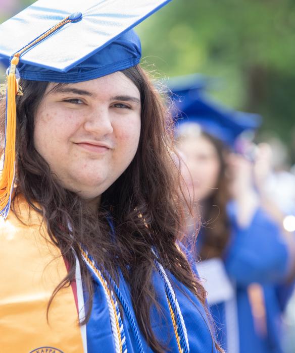 Student in blue cap and gown with yellow tassel and sash looking at the camera smiling.