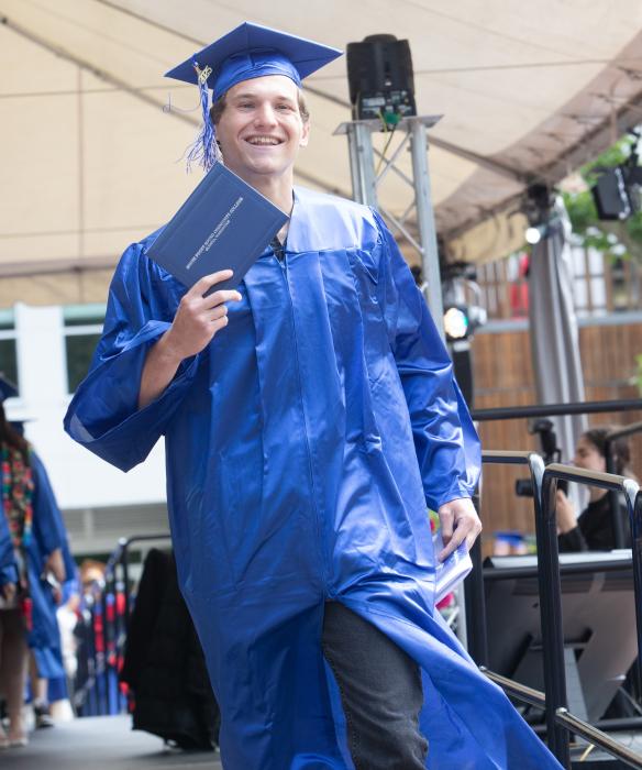 Student in blue graduation cap and gown holding up their diploma smiling.