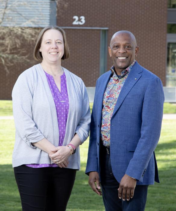 Two people standing in front of a brick building and green grass, facing the camera smiling.