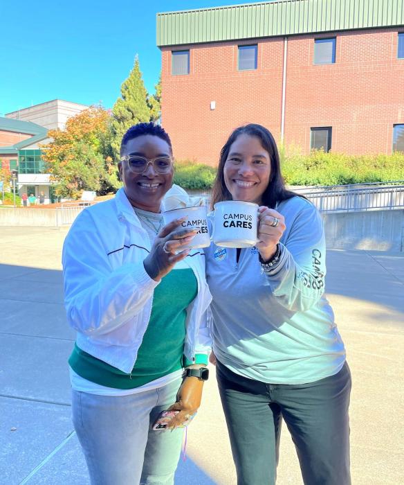 Two people standing outside in front of a brick building smiling. They are both holding mugs that read "Campus Cares".