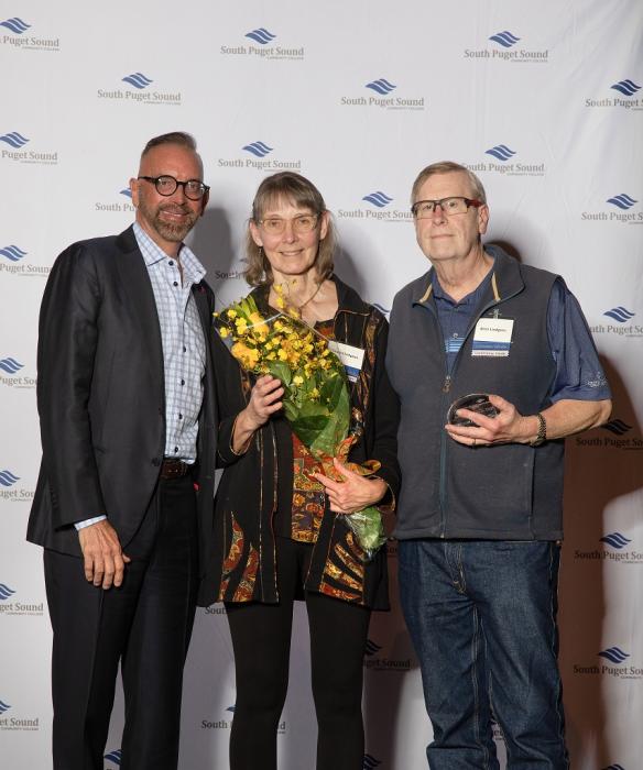 Three people smiling looking at camera, one holding flowers and the other holding a glass trophy.