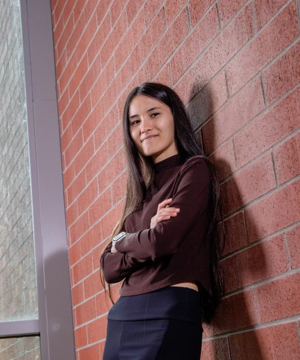 Student leans on brick wall with arms crossed, smiling