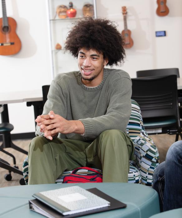 student smiles, sitting in a chair with guitars hanging on the wall behind