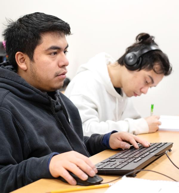 Student in black sweatshirt with hands on a keyboard and mouse, looking toward a desktop computer in a classroom