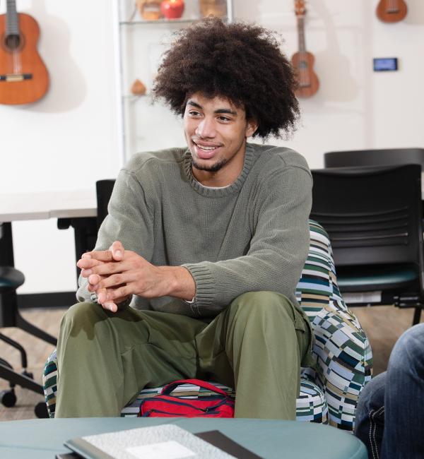 Student sits in front of ottoman, elbows on knees, guitars hanging on wall in background