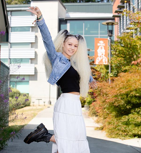 Student stands on one foot with arms in the air, smiling, outside of modern building
