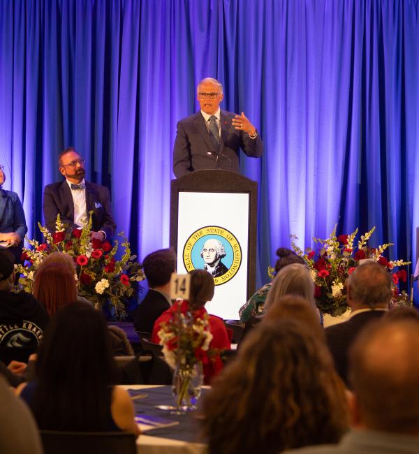 Gov. Inslee at podium speaking to a room, blue curtains in backdrop