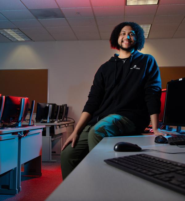 Student leans on table in computer lab with red and blue lights highlighting the space