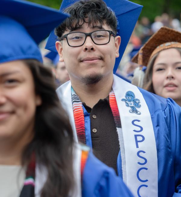 Student in blue graduation Cap and Gown wearing black glasses looks and smirks at the camera.