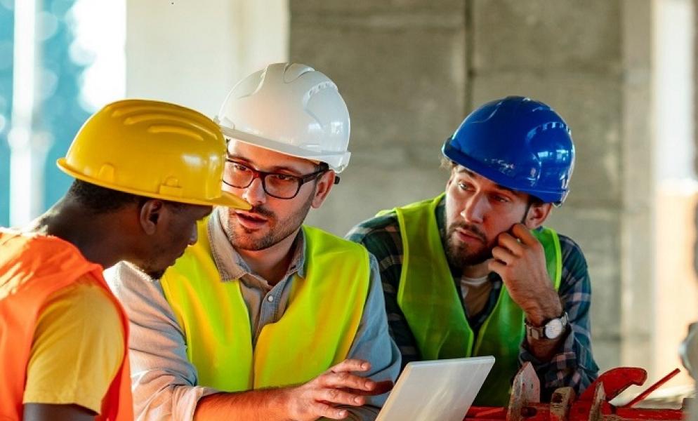 Three people dressed in hard hats and construction wear discussing over a laptop