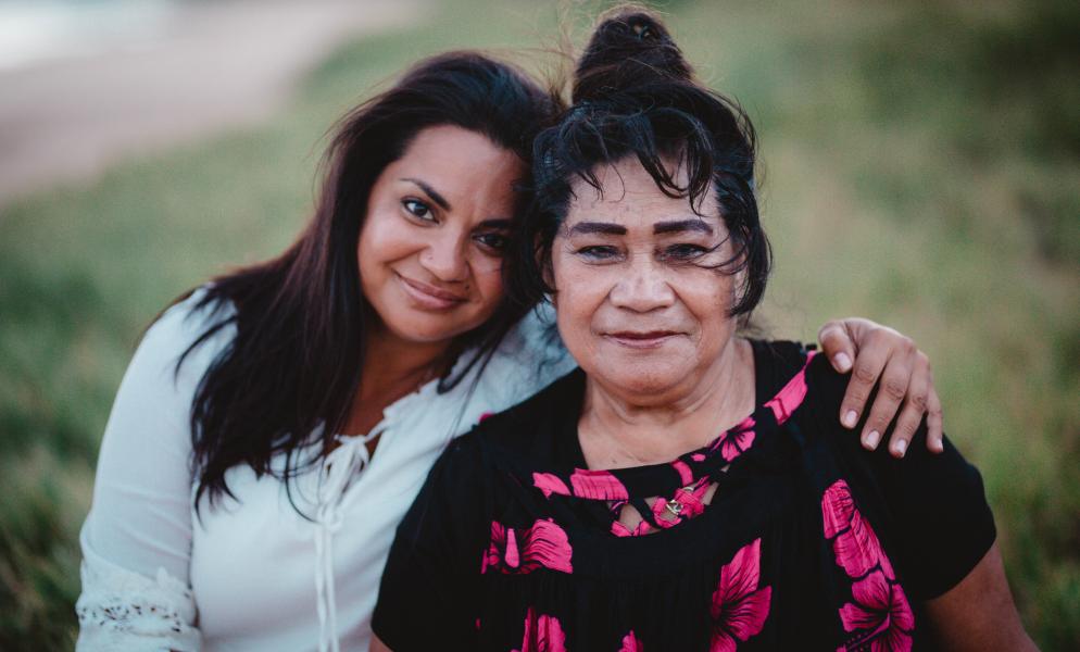Two woman with brown skin and black hair facing the camera smiling. One is wearing a white shirt and the other a black shirt with red print on it.